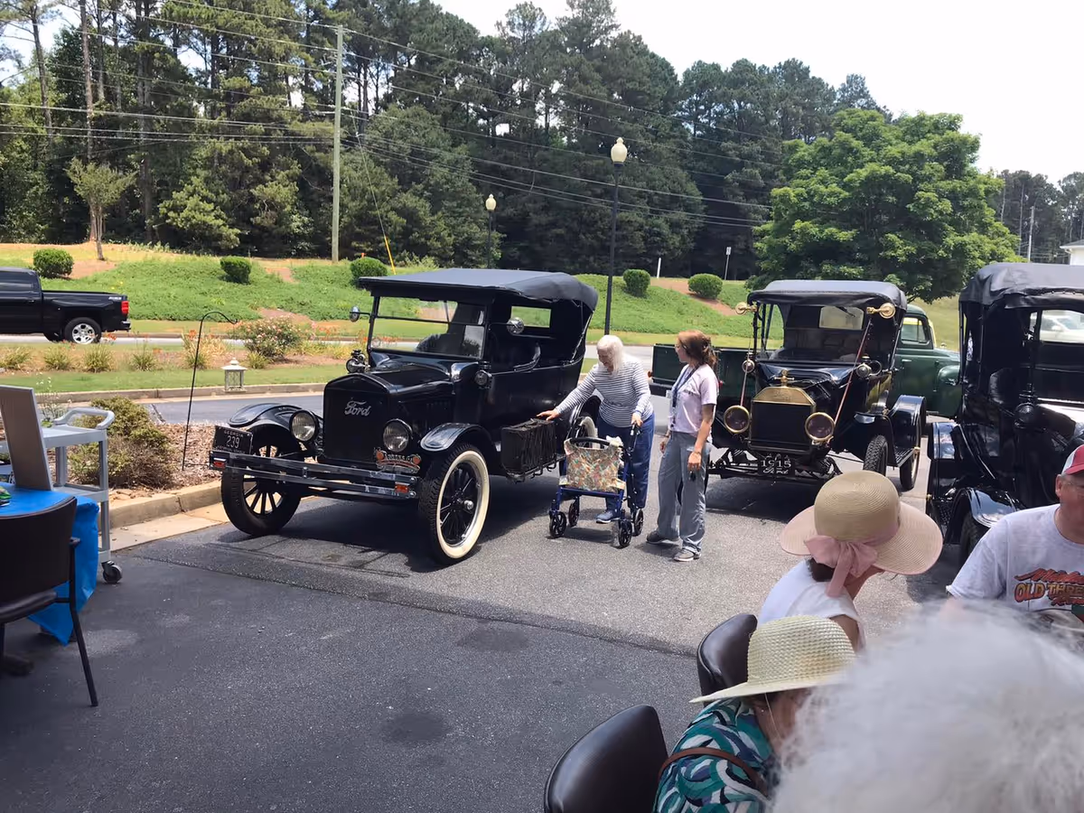 Several people, including elderly residents, stand near vintage Ford cars parked in a facility parking lot with trees in the background.
