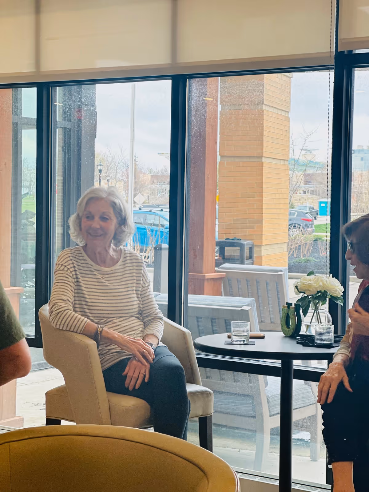 Two elderly women sitting and conversing in a bright indoor seating area with large windows showing an outdoor view with parked cars and brick pillars. A small round table with glasses of water and a vase with white flowers is between them.