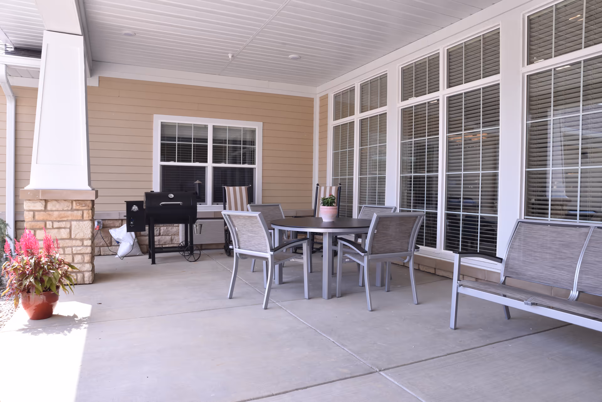 Covered outdoor patio area with a round table surrounded by four chairs, a bench, a potted plant on the table, a barbecue grill, and a potted flowering plant near a stone pillar. The patio is adjacent to a building with beige siding and large windows with white frames and blinds.