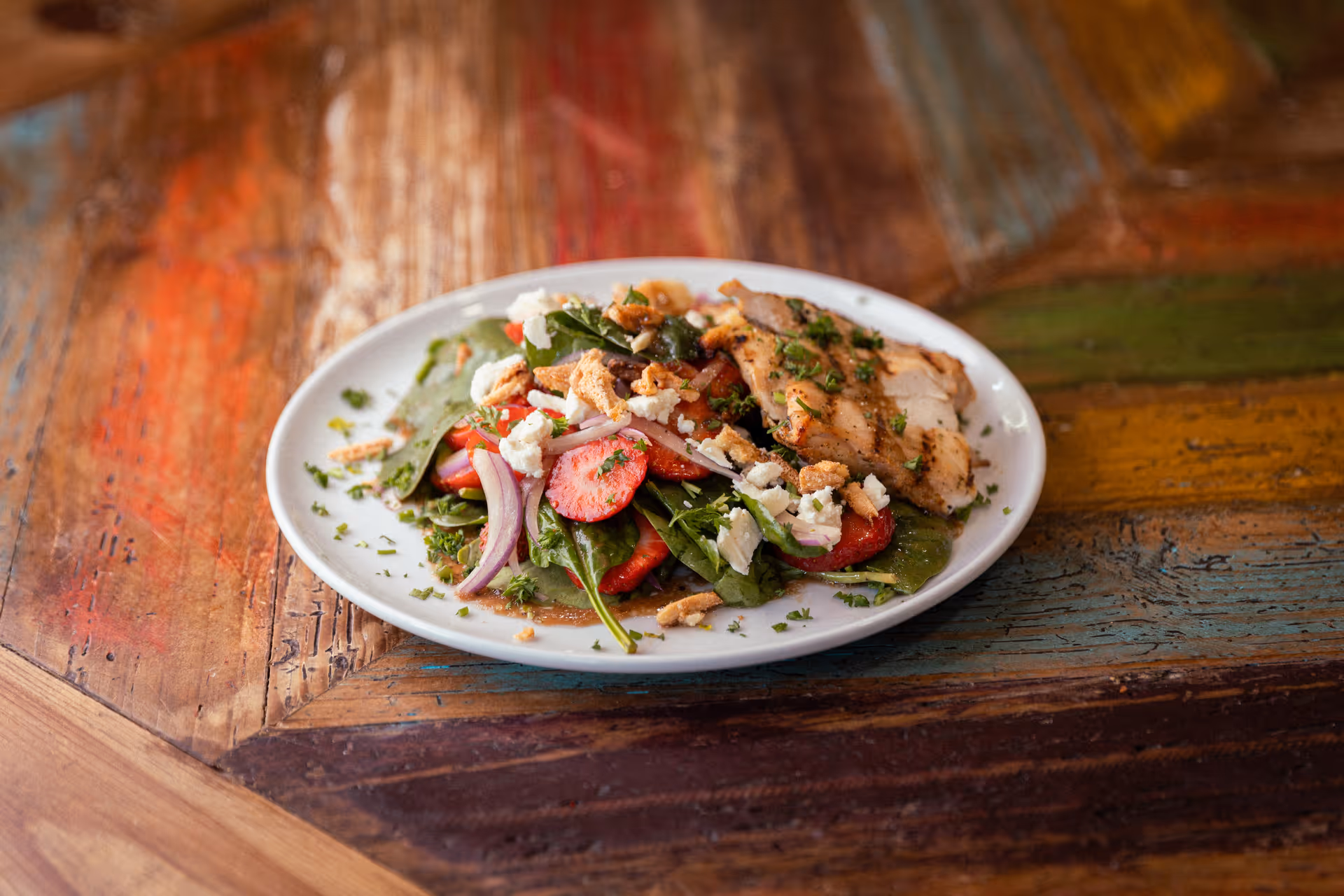 A white plate with a fresh salad consisting of spinach leaves, sliced strawberries, red onion, crumbled cheese, and grilled chicken pieces, garnished with herbs, placed on a colorful wooden table.