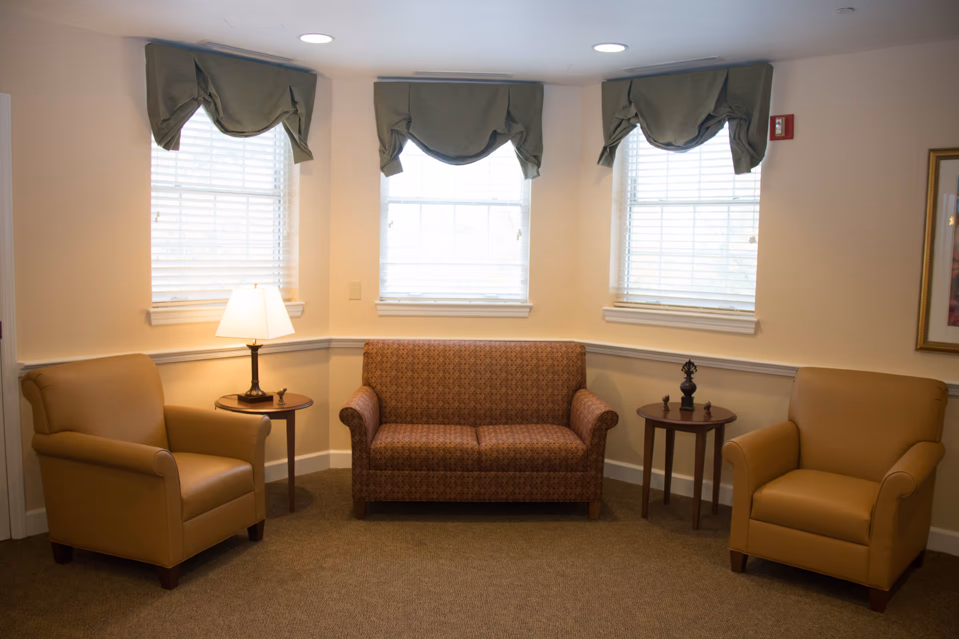A cozy sitting area with two tan armchairs and a patterned loveseat arranged around two small wooden side tables. One table has a lamp, and the other has decorative items. Three windows with white blinds and green valances allow natural light into the room. The walls are light-colored, and there is a framed picture on the right wall.
