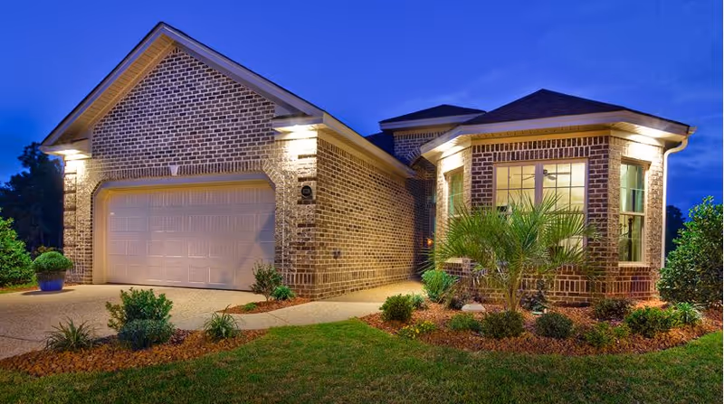 Exterior view of a single-story brick house at dusk with outdoor lighting illuminating the garage door and front windows. The house has a well-maintained lawn and landscaped garden beds with shrubs and small plants.