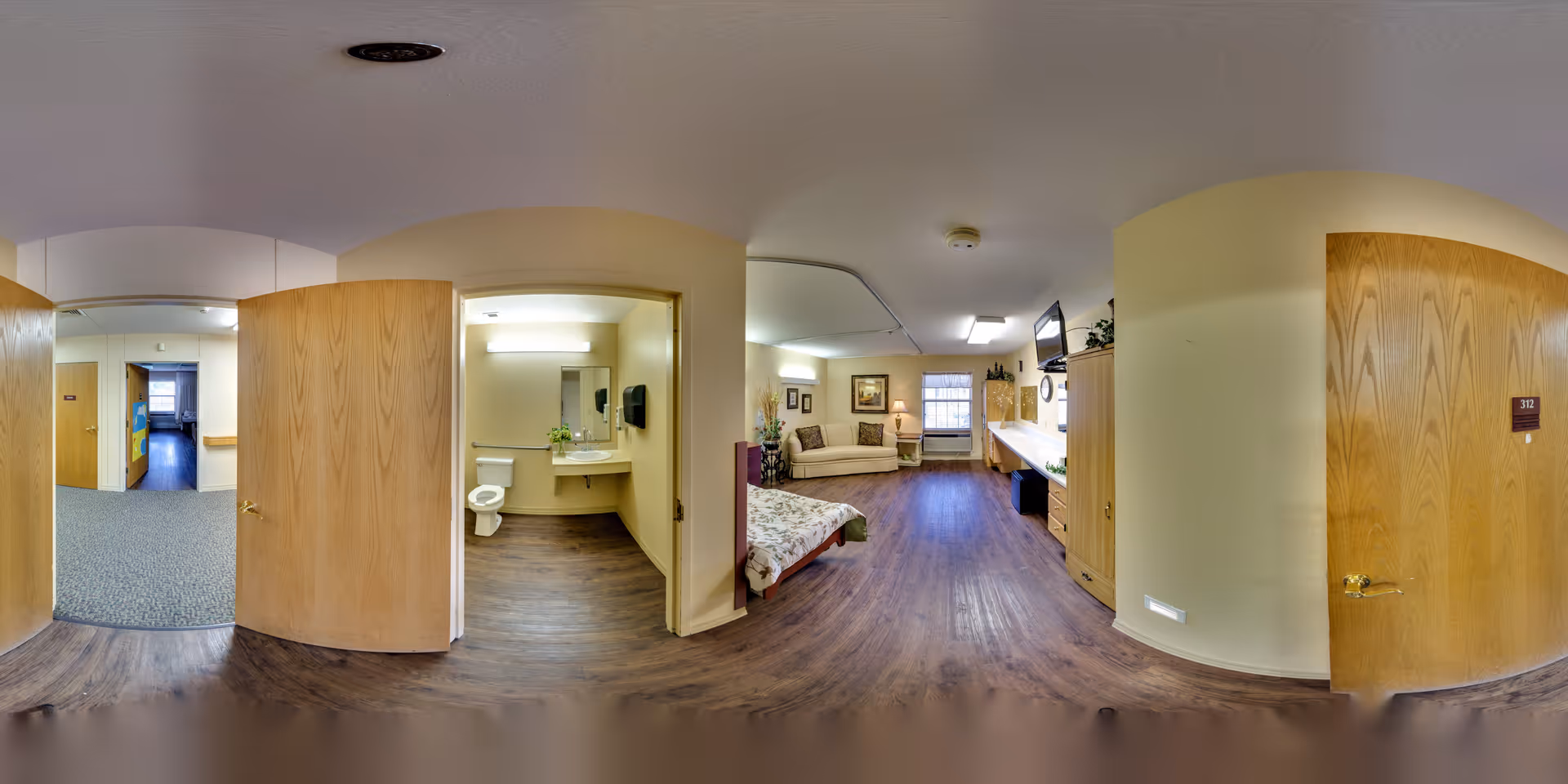Panoramic view of a senior living facility room at Plantation Village Nursing Center showing a bedroom area with a bed, a couch, and a television mounted on the wall. To the left, there is an open door leading to a bathroom with a toilet, sink, and mirror. The room has wooden flooring and light-colored walls with wooden doors.