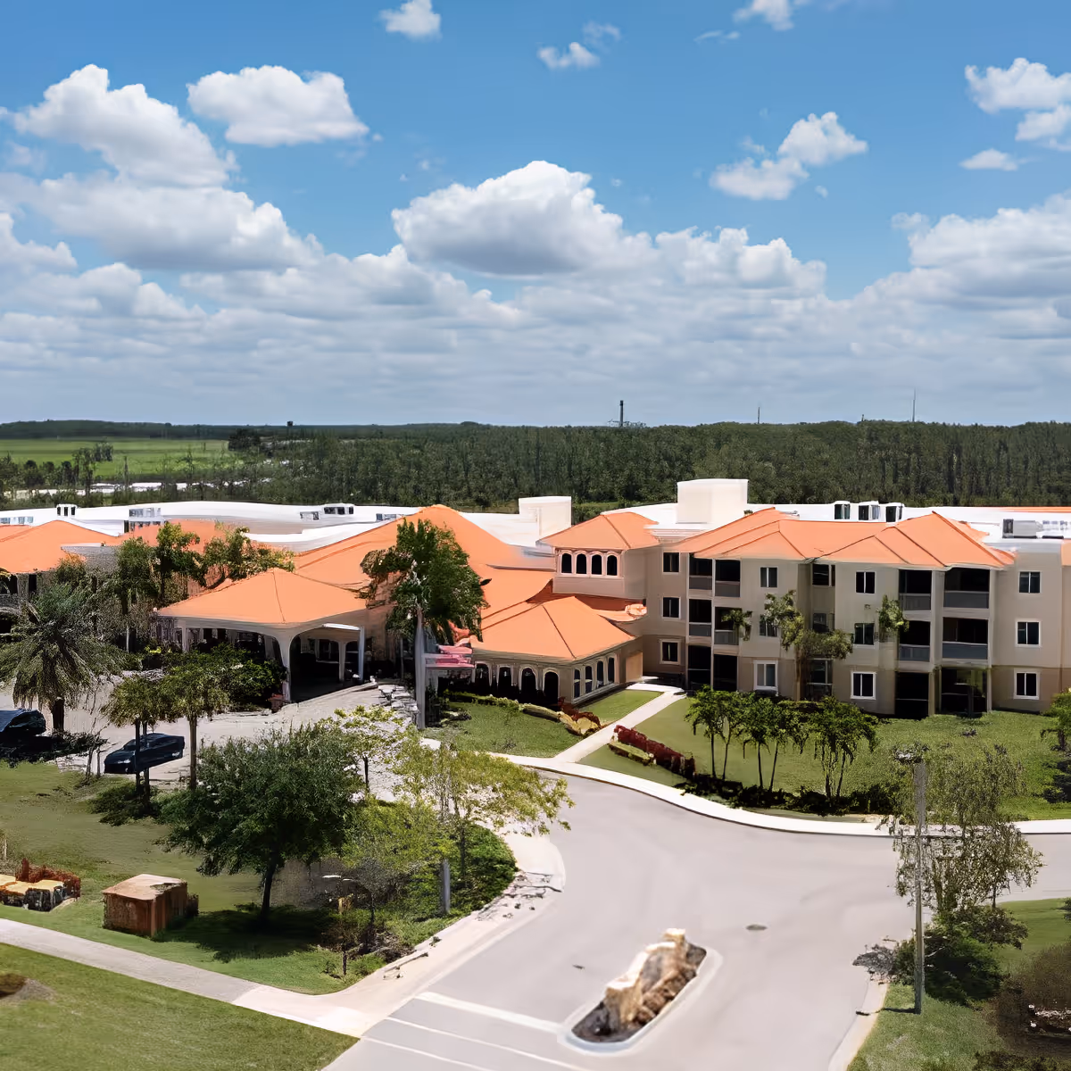 Aerial view of Discovery Village At The Forum, a senior living facility with multiple connected buildings featuring orange roofs, surrounded by greenery and trees under a partly cloudy blue sky.
