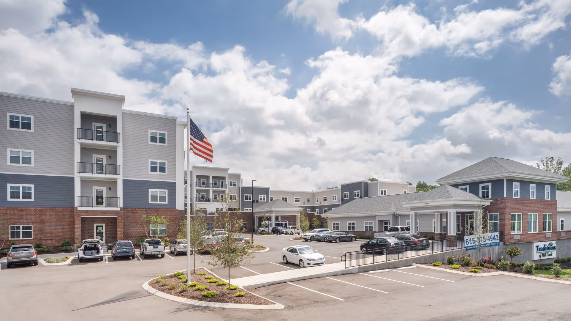 Exterior view of Traditions of Mill Creek senior living facility showing a multi-story building with a combination of brick and siding, a parking lot with several cars, an American flag on a flagpole, and a partly cloudy sky.