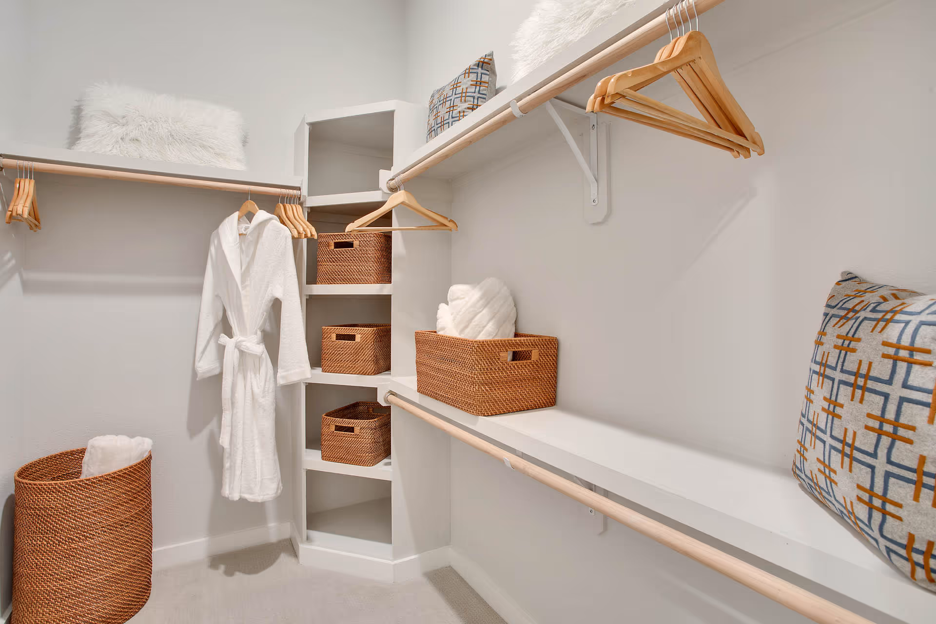 A spacious walk-in closet with white walls and carpeted floor. The closet features wooden hanging rods with several wooden hangers, a white bathrobe hanging on one rod, and multiple woven baskets placed on built-in shelves and a bench. There are also decorative pillows and a fluffy white cushion on the top shelf.