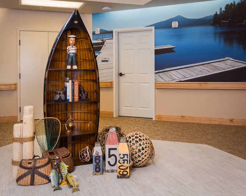 An indoor space with a wooden boat-shaped bookshelf containing books and decorative items, including a sailor figurine. In front of the bookshelf are various nautical-themed decorations such as fishing nets, wooden buoys, and wicker baskets. The background features a large wall mural of a serene lake with docks and mountains.