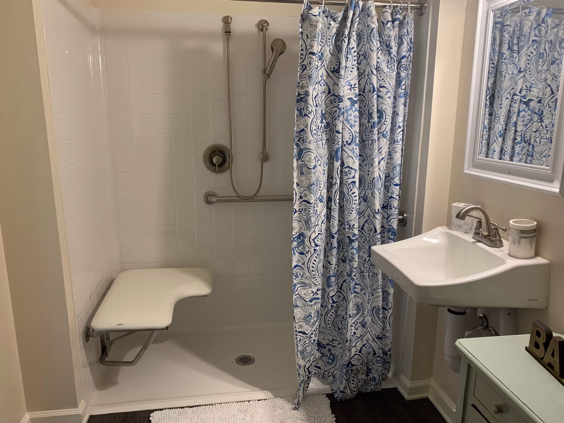 A bathroom with a walk-in shower featuring a fold-down seat and a handheld showerhead. The shower has white tiled walls and a blue and white patterned shower curtain. Next to the shower is a white sink with a silver faucet, a mirror above it, and a small green cabinet with a decorative item on top.