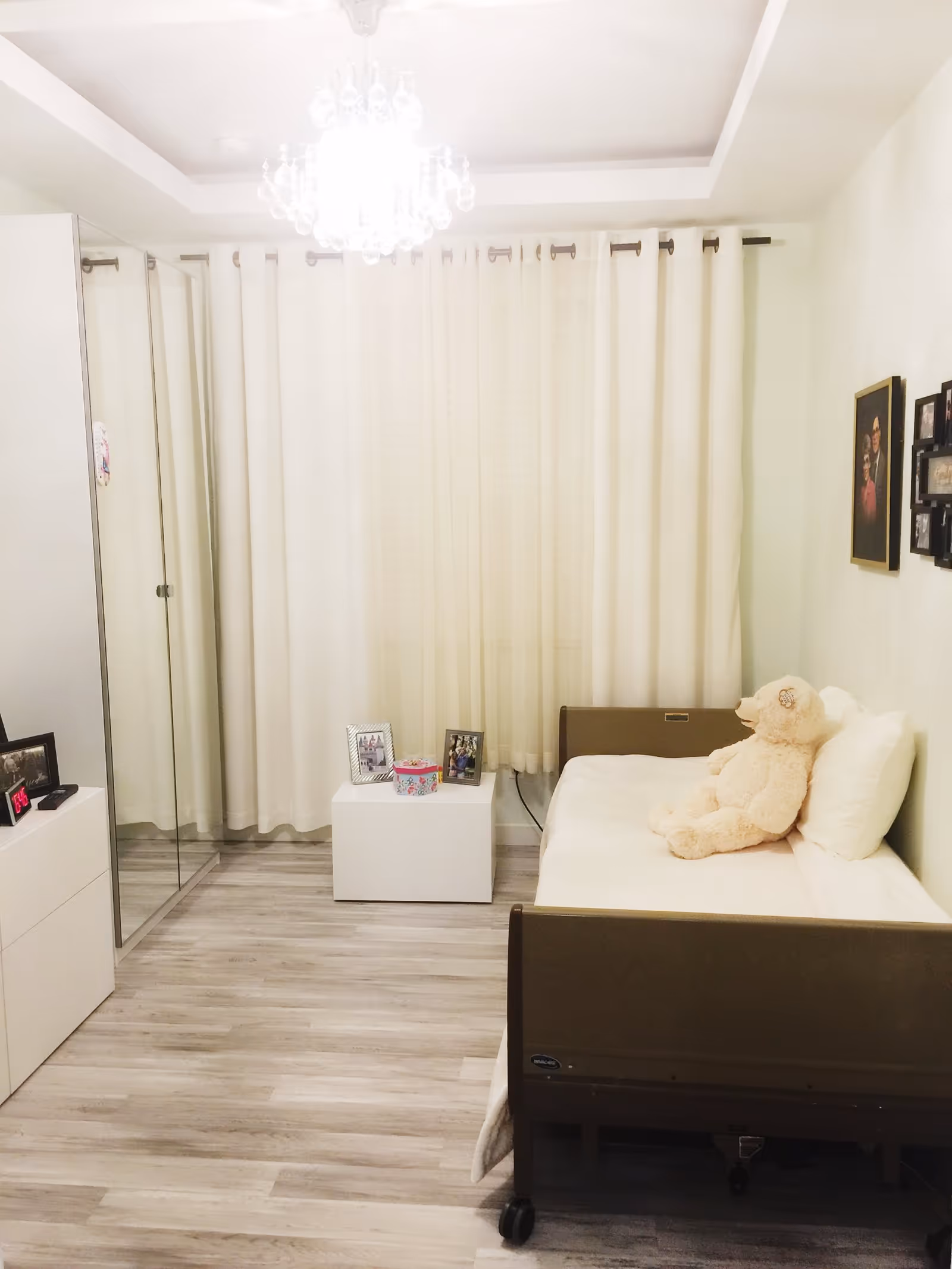 A small, neatly arranged bedroom with light-colored walls and wood flooring. The room features a single bed with a white pillow and blanket, and a large teddy bear sitting on the bed. There is a white nightstand next to the bed with framed photos and a decorative box. A mirrored wardrobe is on the left side of the room, and a chandelier hangs from the ceiling. The window is covered with white curtains.