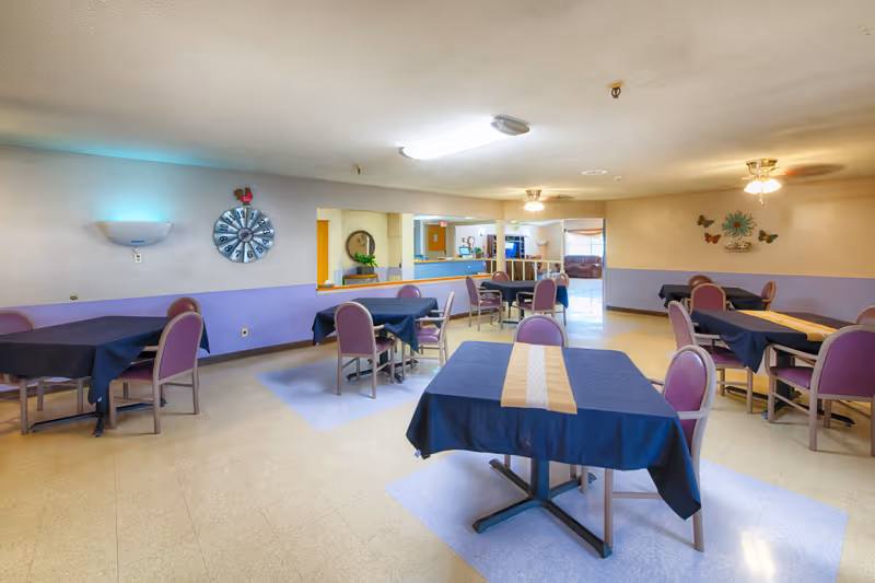 Communal dining room with several tables covered in dark tablecloths and mauve chairs under ceiling lights.