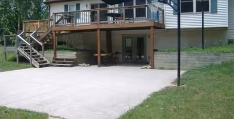 View of the back exterior of a house with a raised wooden deck and stairs leading down to a concrete patio area. The house has white siding and a grassy lawn surrounding the patio.