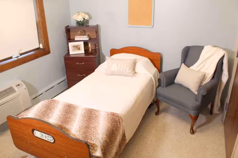 A small, cozy bedroom with a single wooden bed covered in white bedding and a brown patterned blanket at the foot. There is a beige pillow with the word 'home' on it. Next to the bed is a dark wooden nightstand with drawers, decorated with a vase of white flowers, a framed photo, and some books. A gray upholstered armchair with a beige pillow and a white throw blanket is positioned beside the bed. The room has light-colored walls, a window with a wooden frame, and a wall-mounted air conditioning unit below the window.