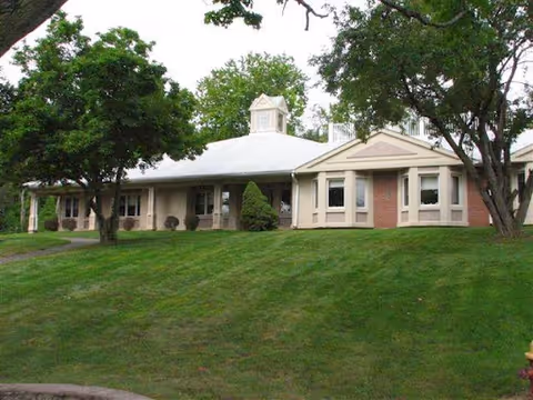 Exterior view of a single-story building with a light-colored roof and brick walls, surrounded by green grass and trees under a cloudy sky.