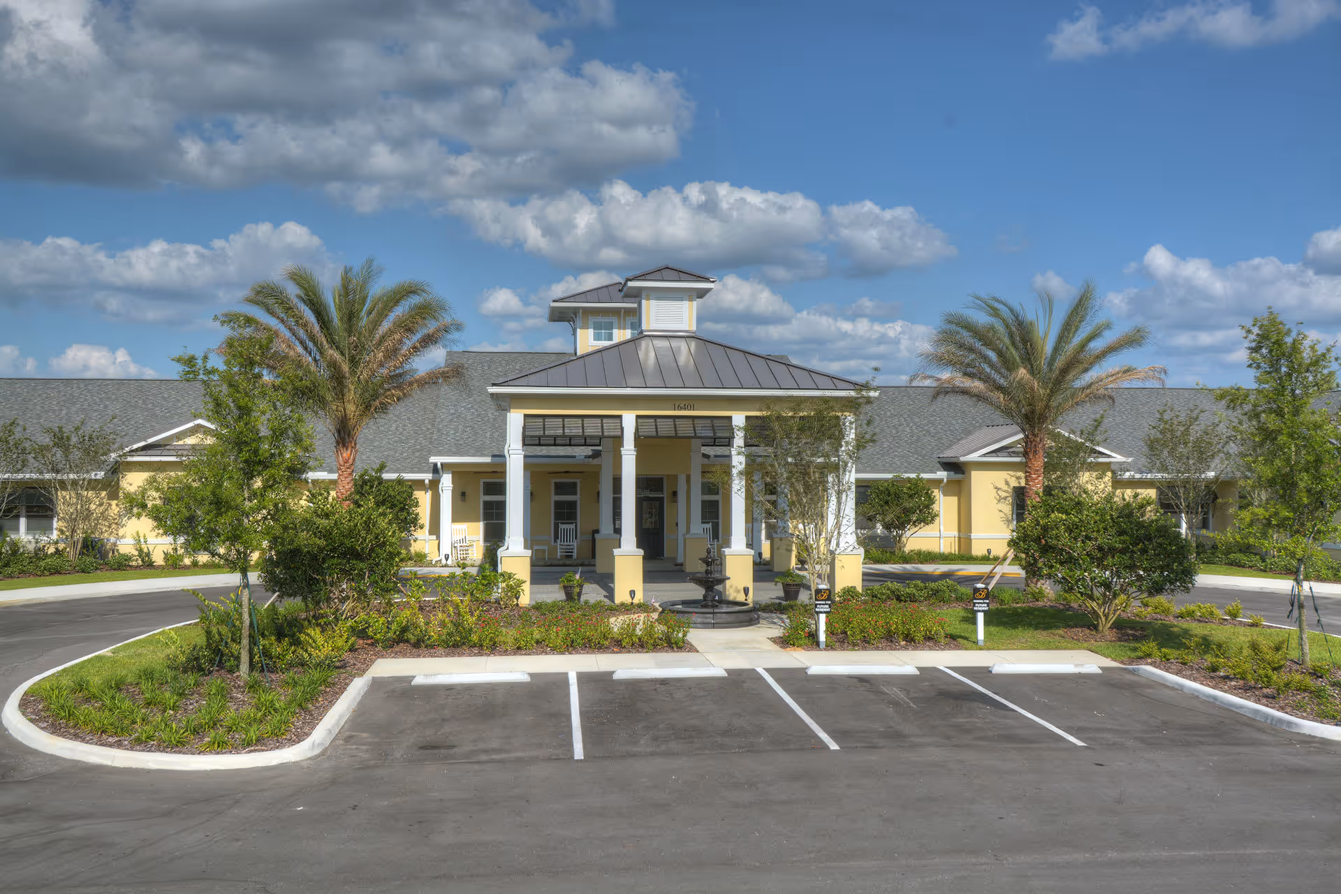 Front entrance of a single-story yellow senior living facility with a covered portico, palm trees, landscaped beds, and empty parking spaces under a blue sky.