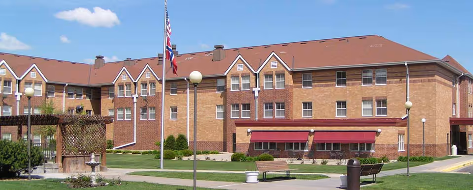 Brick three-story senior living building with a red roof, flagpole, benches and landscaped lawn in front.