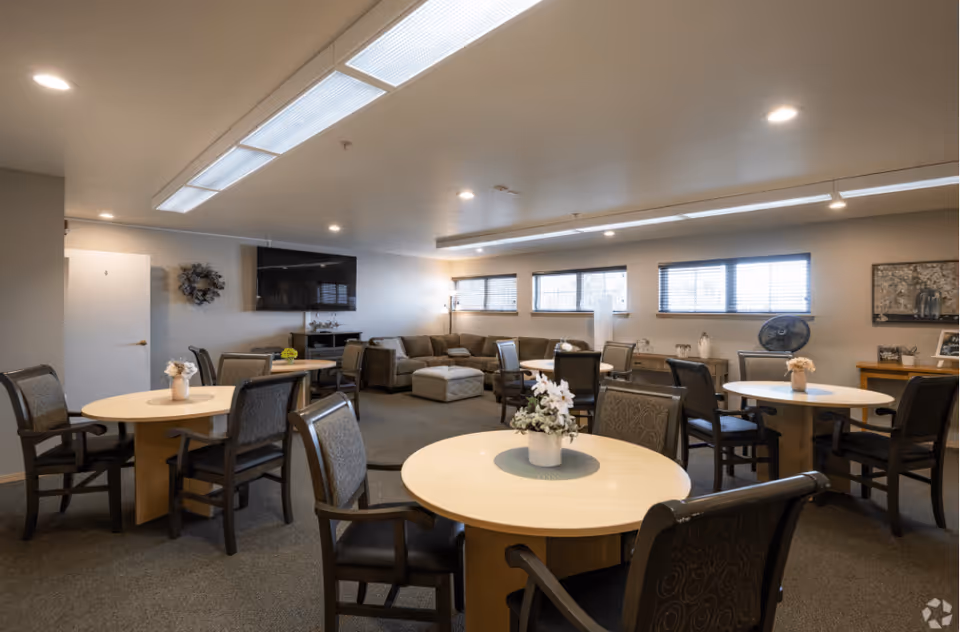A spacious common area with multiple round tables surrounded by chairs, each table decorated with a small flower arrangement. In the background, there is a large sectional sofa with an ottoman, a flat-screen TV mounted on the wall, and several windows letting in natural light. The room has a neutral color palette and overhead fluorescent lighting.