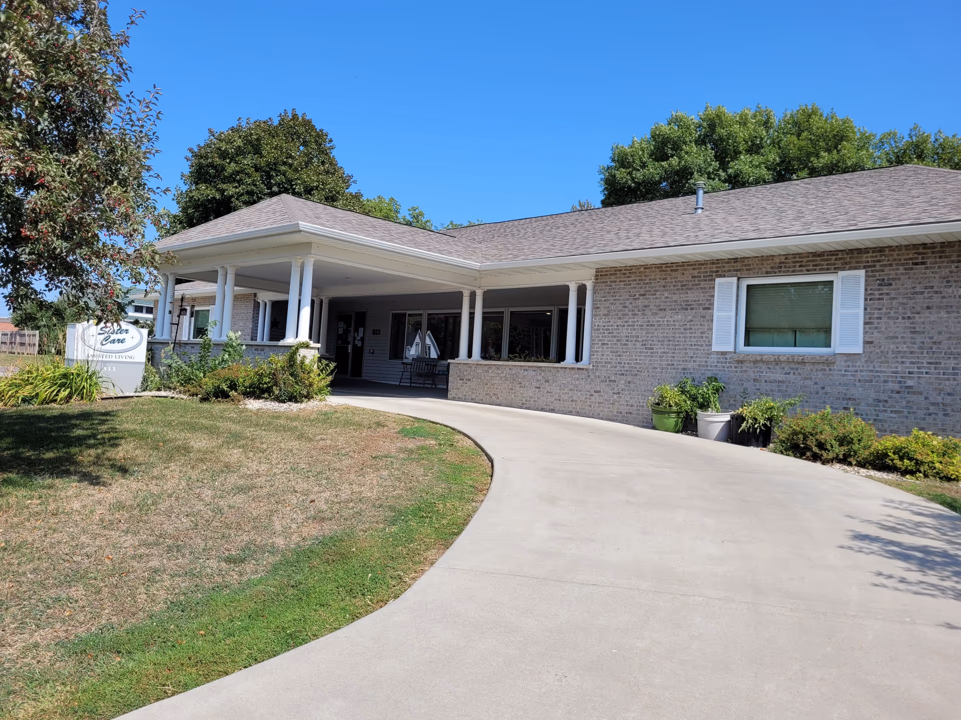 Exterior view of a single-story brick building with a covered entrance supported by white columns. There is a curved concrete driveway leading to the entrance. A sign near the entrance reads 'Sister Care Assisted Living'. The surrounding area has some grass, bushes, and trees under a clear blue sky.