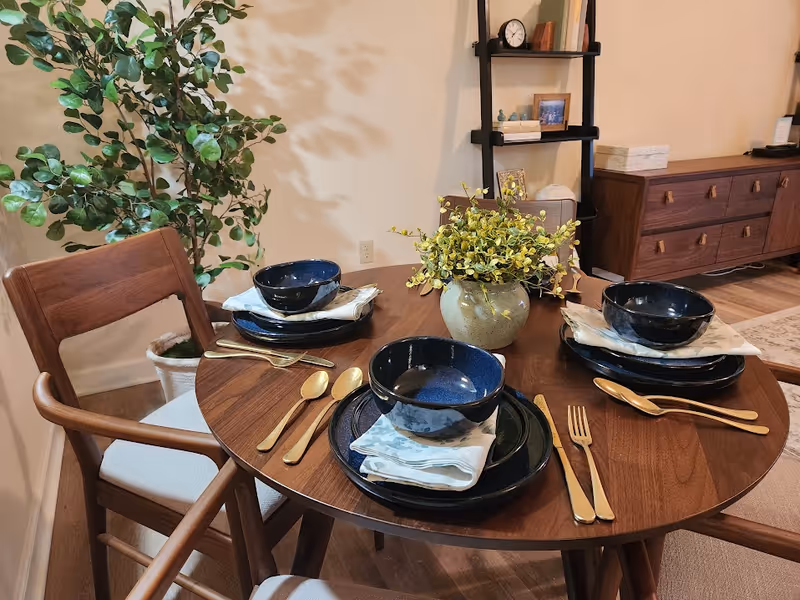 A round wooden dining table set for three with dark blue bowls, plates, and gold-colored cutlery. Each place setting has a folded napkin under the bowl. A vase with yellow-green foliage is in the center of the table. The background includes a potted plant, a black ladder shelf with decorative items, and a wooden sideboard.