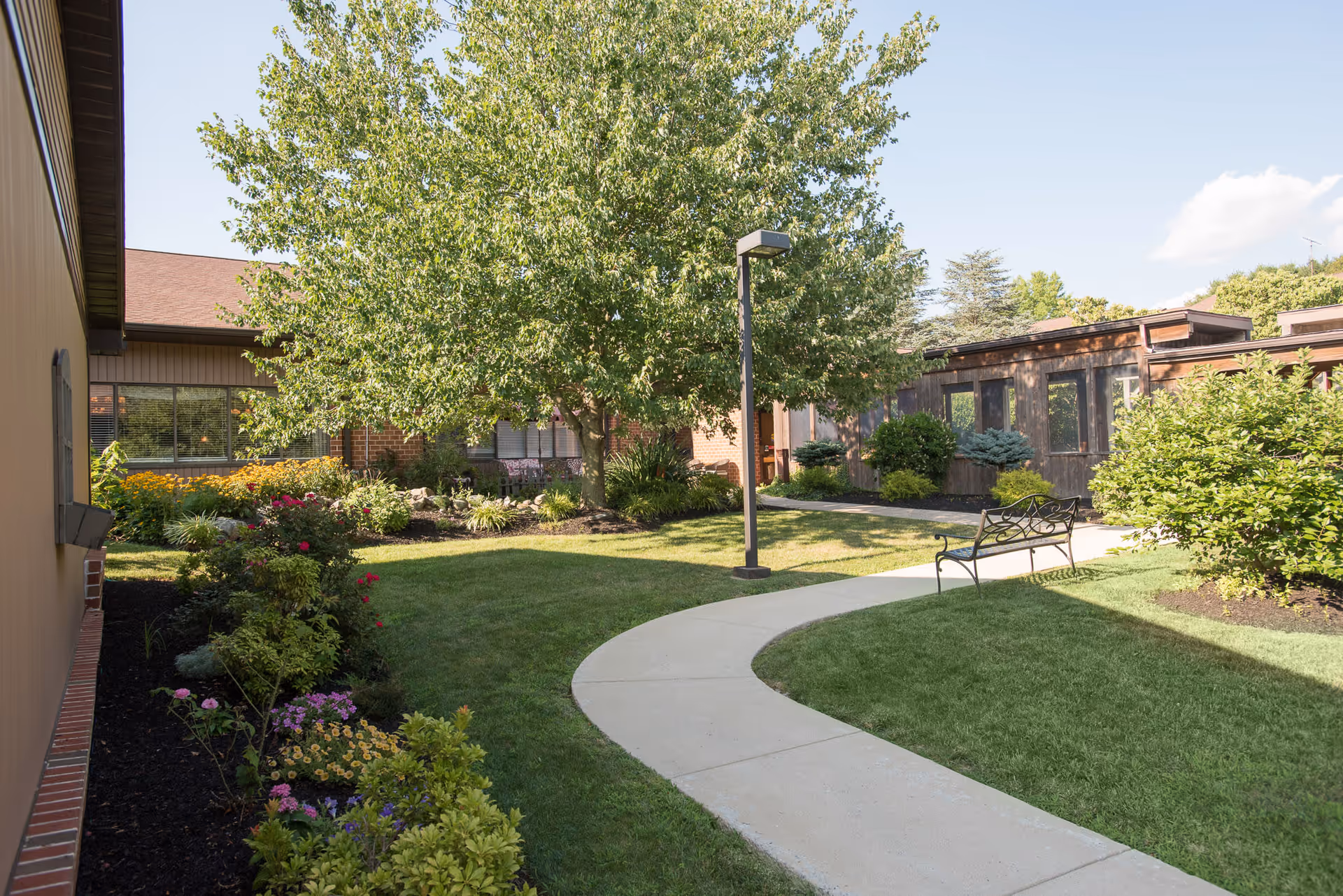 A sunny outdoor courtyard at Heatherwood Retirement Community featuring a curved concrete pathway, green grass, a variety of flowering plants and shrubs, a large leafy tree, a metal bench, and surrounding buildings with windows.