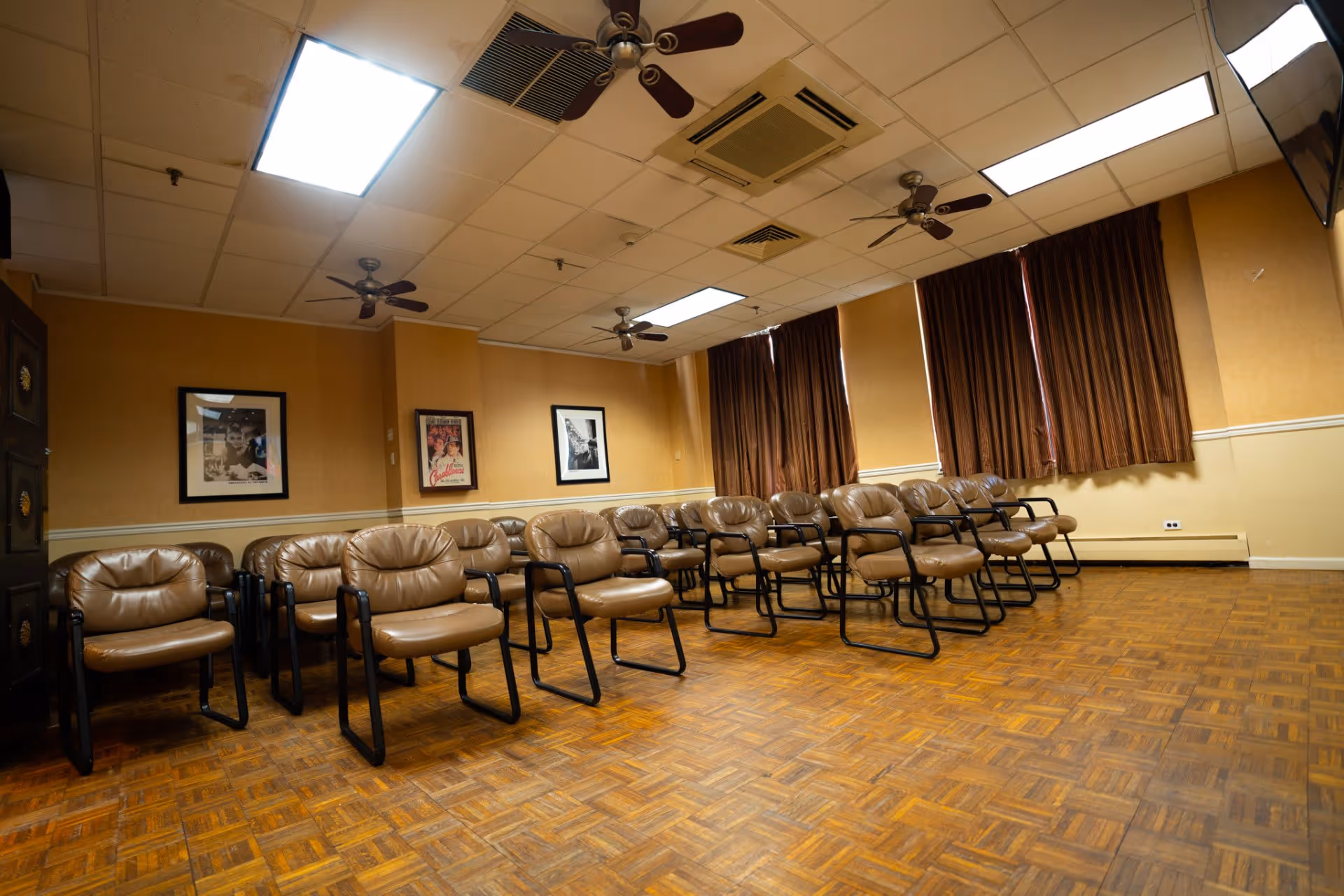 Empty activity/meeting room with rows of brown leather chairs on a parquet floor, ceiling fans, and closed curtains.