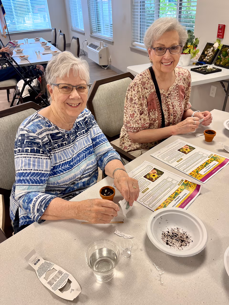 Two elderly women sitting at a table in a well-lit room, smiling at the camera while engaging in a planting activity. They have small pots, soil, and instruction sheets in front of them. The room has large windows with blinds and several chairs and tables in the background.