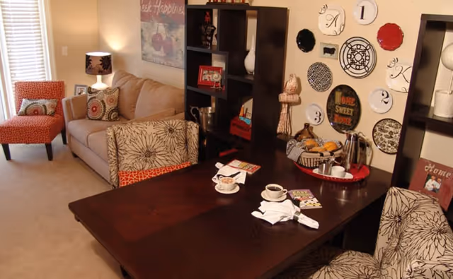 A cozy living room area with a beige sofa, patterned armchairs, and a dark wooden table set with coffee cups, napkins, and a tray of fruit and coffee items. The wall behind the table is decorated with various decorative plates and a 'Home Sweet Home' sign. Shelves with decorative items and framed pictures are also visible.