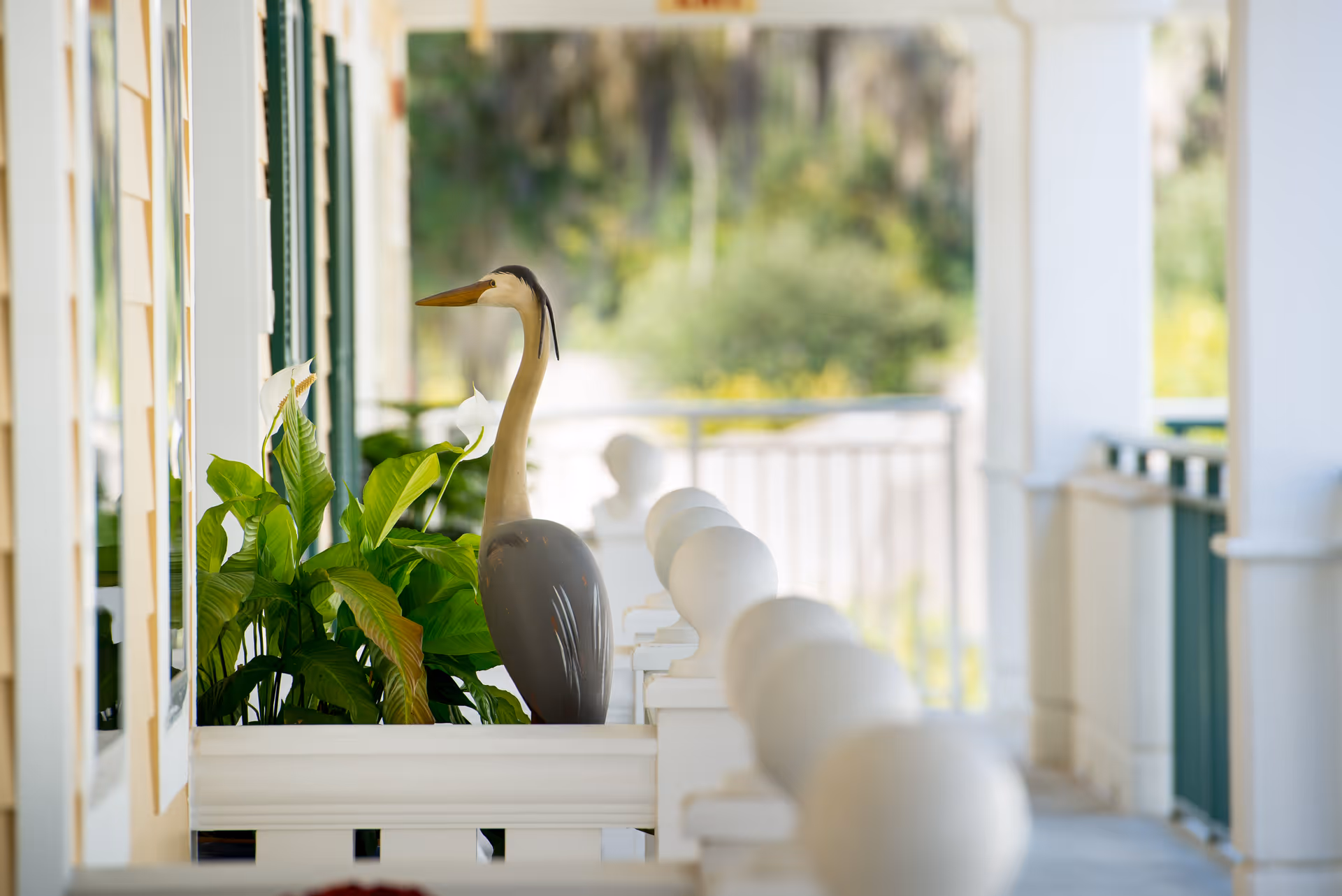 Covered porch with a planter of leafy plants and a decorative heron statue along a white railing overlooking blurred greenery.