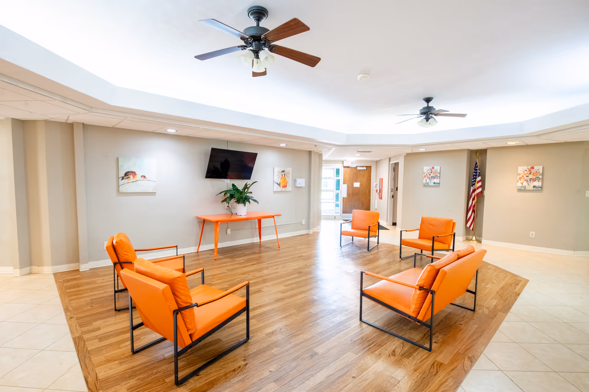 A bright and spacious common area with a wooden floor section surrounded by beige tiled flooring. The room features five orange cushioned chairs with black metal frames arranged in a semi-circle. There is an orange table against the wall with a potted plant on it, and a flat-screen TV mounted above the table. The walls are light gray with colorful artwork hanging on them. Two ceiling fans with lights are mounted on the white ceiling. An American flag stands in the corner near a doorway.
