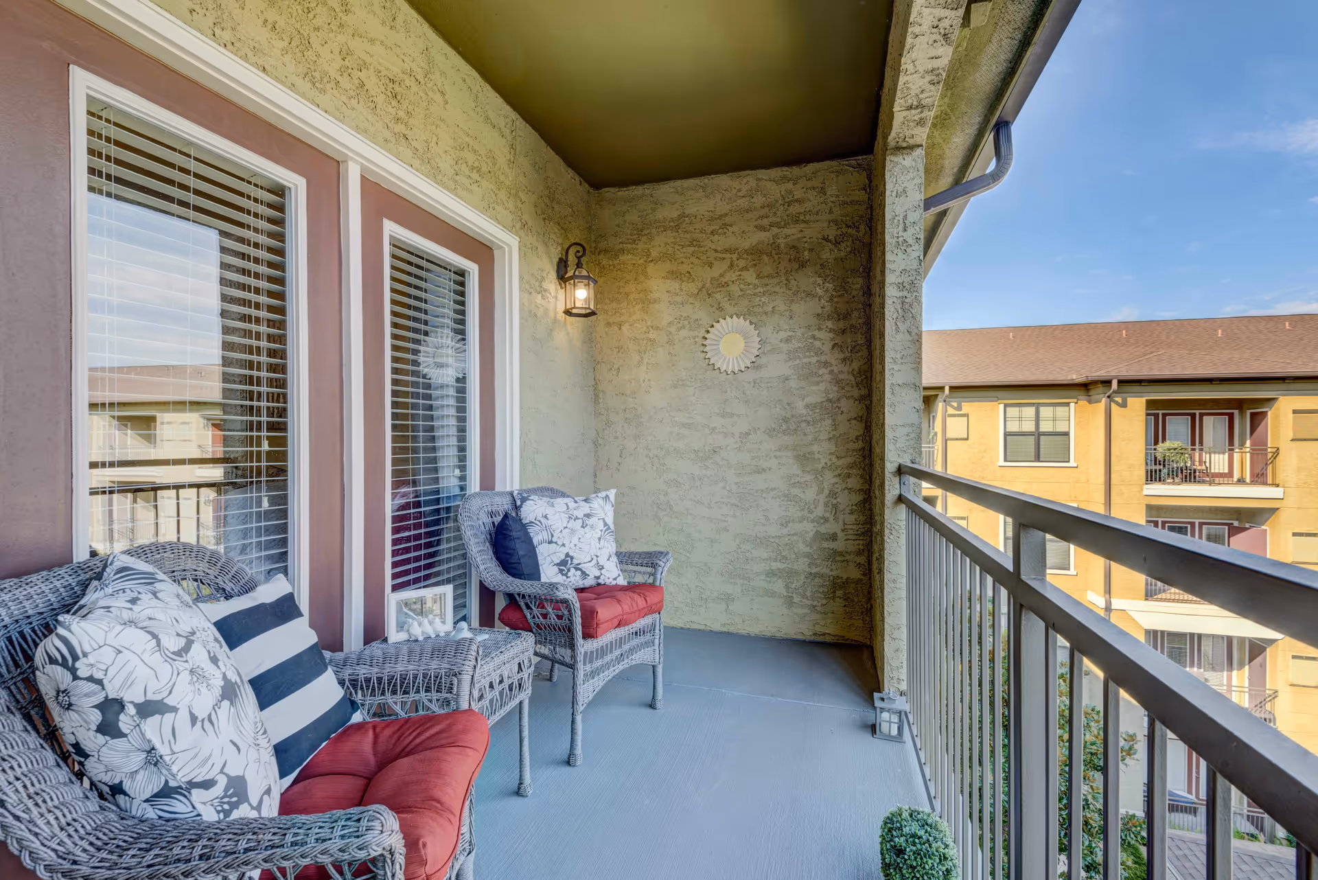Covered balcony with wicker chairs and cushions, a small table, and a railing overlooking neighboring apartment buildings.
