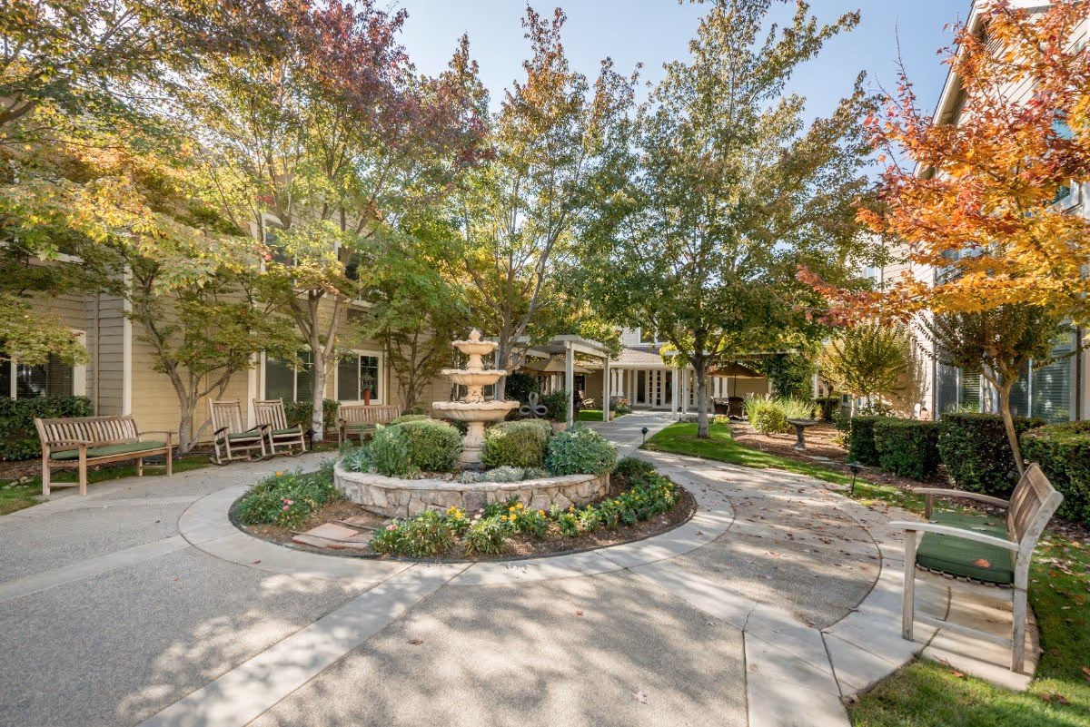 Outdoor courtyard area at The Grove Senior Living featuring a circular stone fountain surrounded by greenery and flowers. There are several wooden benches along the paved pathways, with trees showing autumn foliage and buildings in the background.