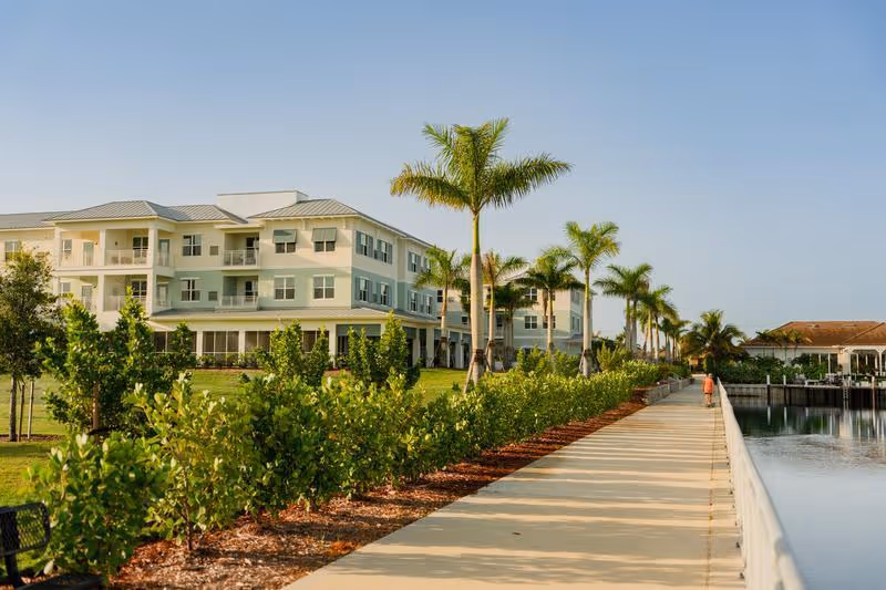 A large, light-colored multi-story building with balconies, surrounded by palm trees and greenery, next to a wide paved walkway along a body of water under a clear blue sky.
