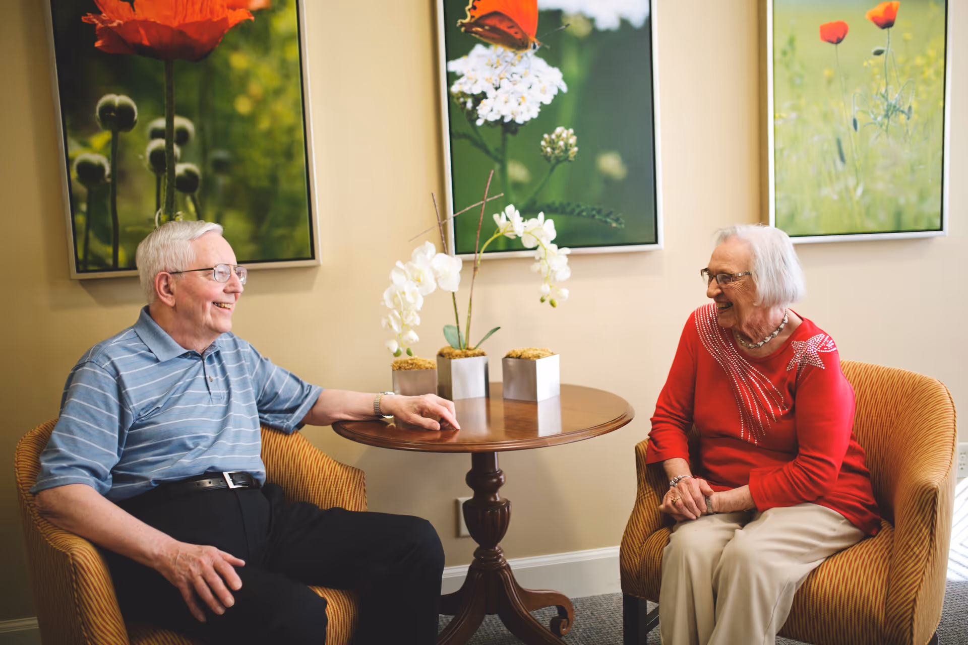 An elderly man and woman sitting in armchairs facing each other and smiling in a cozy room with a round wooden table between them holding three small potted white orchid plants. Behind them are three framed pictures of flowers on the wall.