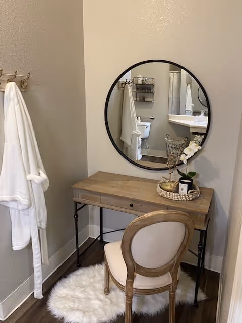 A small vanity area with a wooden desk and a cushioned chair on a white fluffy rug. A round mirror hangs on the wall above the desk, reflecting a bathroom with a toilet and sink. Two white robes hang on hooks on the left wall.