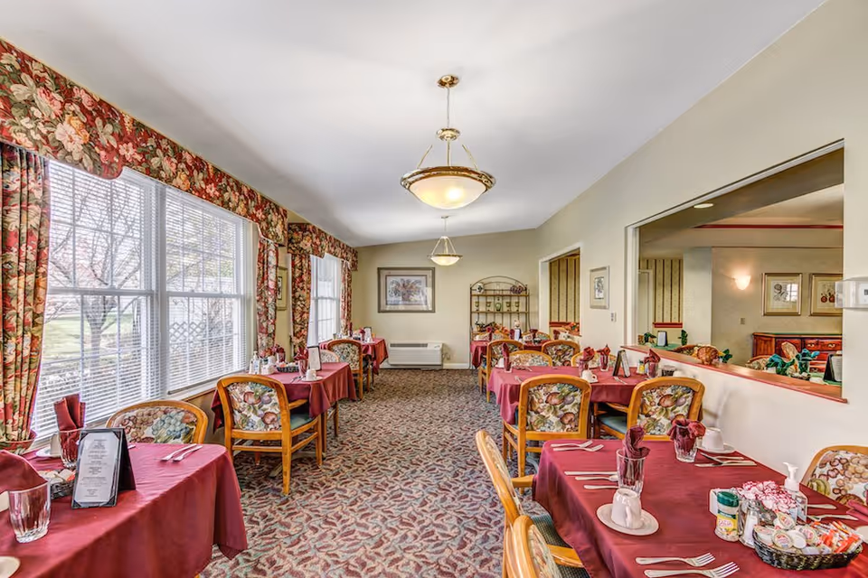 A dining room with several tables covered in burgundy tablecloths, each set with utensils, glasses, napkins, and condiments. The chairs have floral upholstery. Large windows with floral curtains let in natural light, and the room has patterned carpet and beige walls with framed artwork. Two hanging light fixtures illuminate the space.