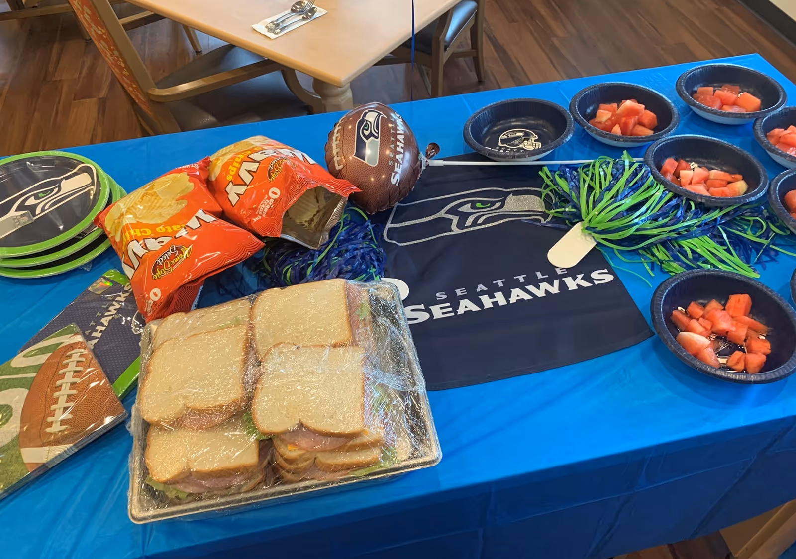 A table covered with a blue Seattle Seahawks tablecloth and decorated with Seahawks-themed plates, napkins, and a small football balloon. On the table are trays of sandwiches wrapped in plastic, bags of Lay's potato chips, bowls of cut watermelon, and green and blue pom-poms.