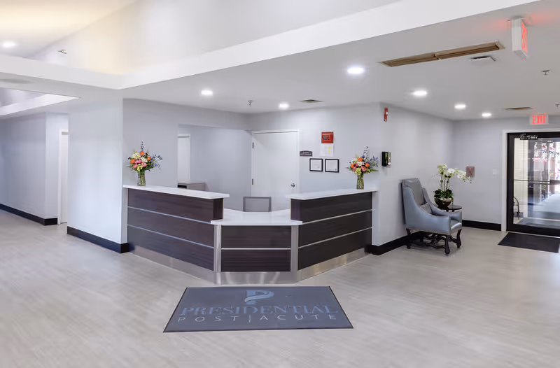 Reception area of Presidential Post Acute facility with a modern dark wood and white reception desk, two flower arrangements on the desk, a gray armchair with a small table and plant next to it, light-colored flooring, and an exit door with glass panels on the right side.