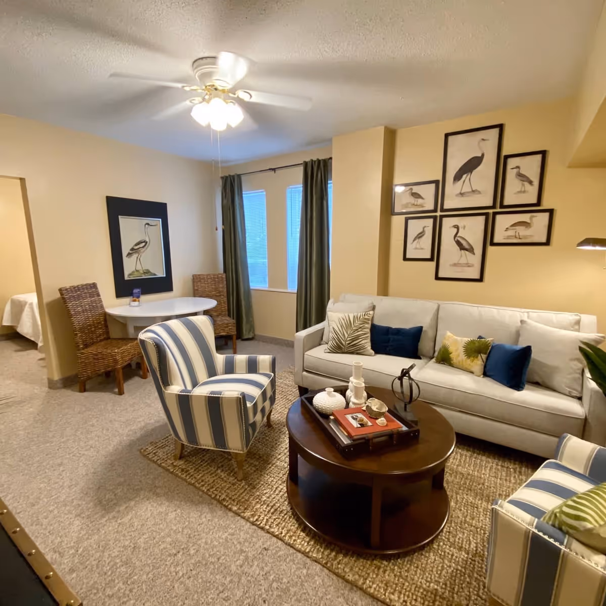 A cozy living room with a beige sofa adorned with decorative pillows, two striped armchairs, and a round wooden coffee table with decorative items. The walls are painted light yellow and decorated with framed bird artwork. A small round dining table with two wicker chairs is positioned near a window with green curtains. A ceiling fan with lights is mounted on the ceiling. A glimpse of a bedroom with a bed is visible through an open doorway.