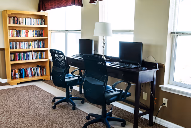 A small computer workstation area with two black office chairs in front of a dark wooden desk holding two desktop computers and a lamp, next to three large windows with white blinds. To the left, there is a wooden bookshelf filled with books. The floor is carpeted with a patterned brown carpet.
