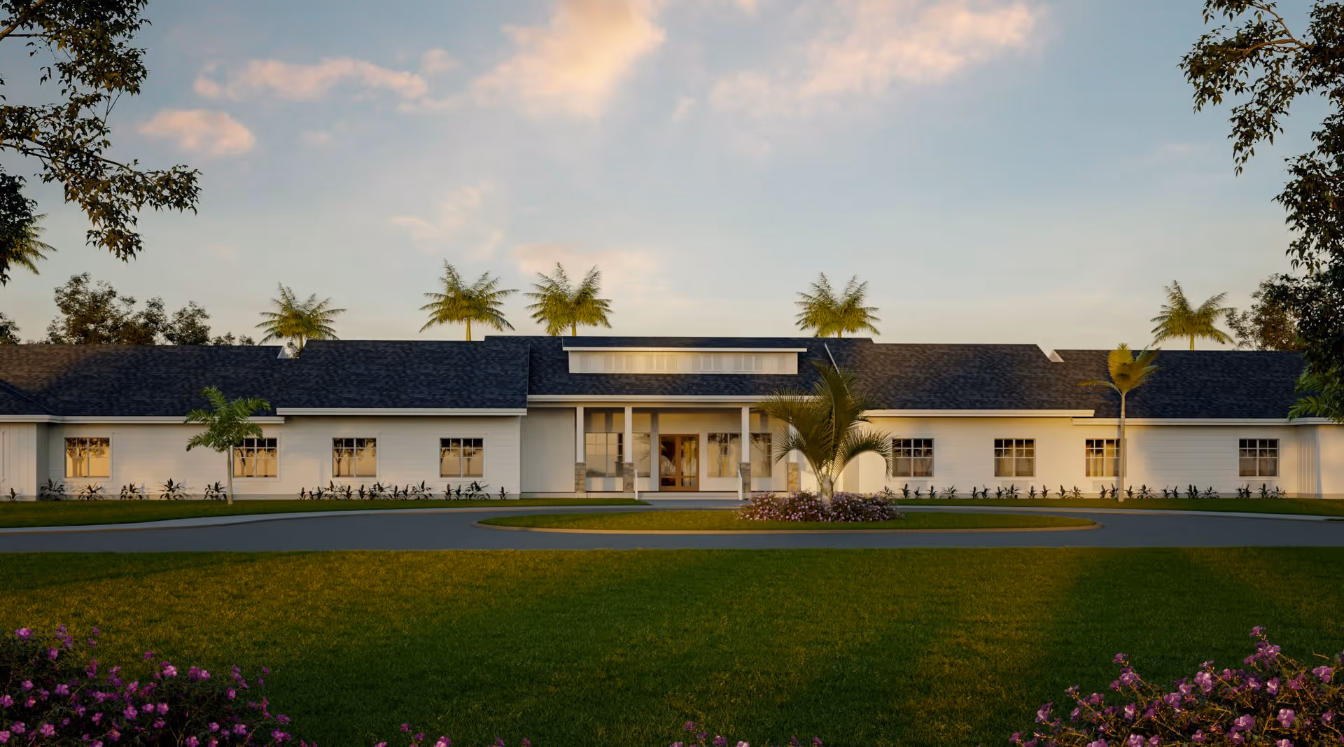 Exterior view of a single-story senior living facility building with a dark shingled roof, white walls, and multiple windows. The building is surrounded by green grass, palm trees, and flowering plants under a partly cloudy sky during sunset or sunrise.
