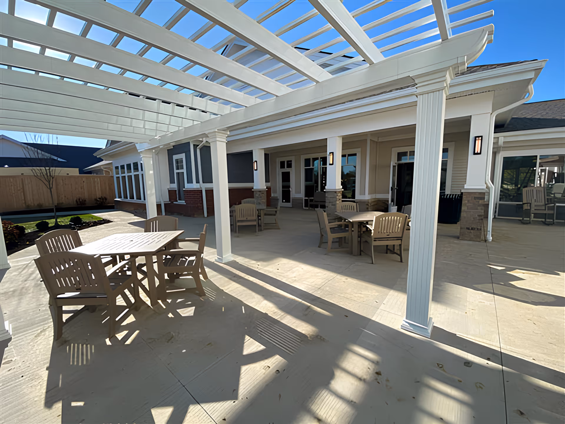 Outdoor patio area at The Ashford of Grove City with several tables and chairs under a white pergola. The patio is adjacent to the building with large windows and doors, and there is a wooden fence and some landscaping visible in the background under a clear blue sky.