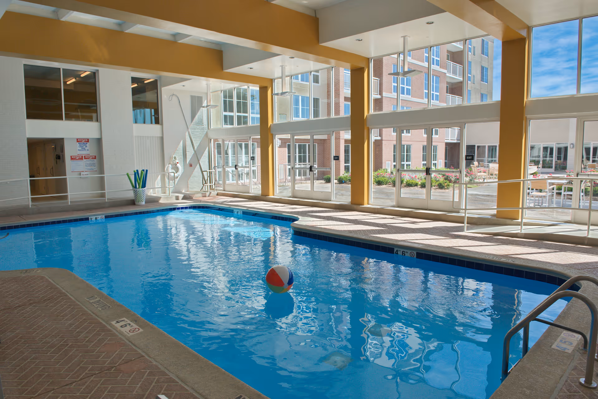 Indoor swimming pool with clear blue water and a colorful beach ball floating on the surface. Large windows let in natural light and offer a view of an outdoor patio area and a multi-story brick building. Poolside area has tiled flooring and a metal pool ladder.