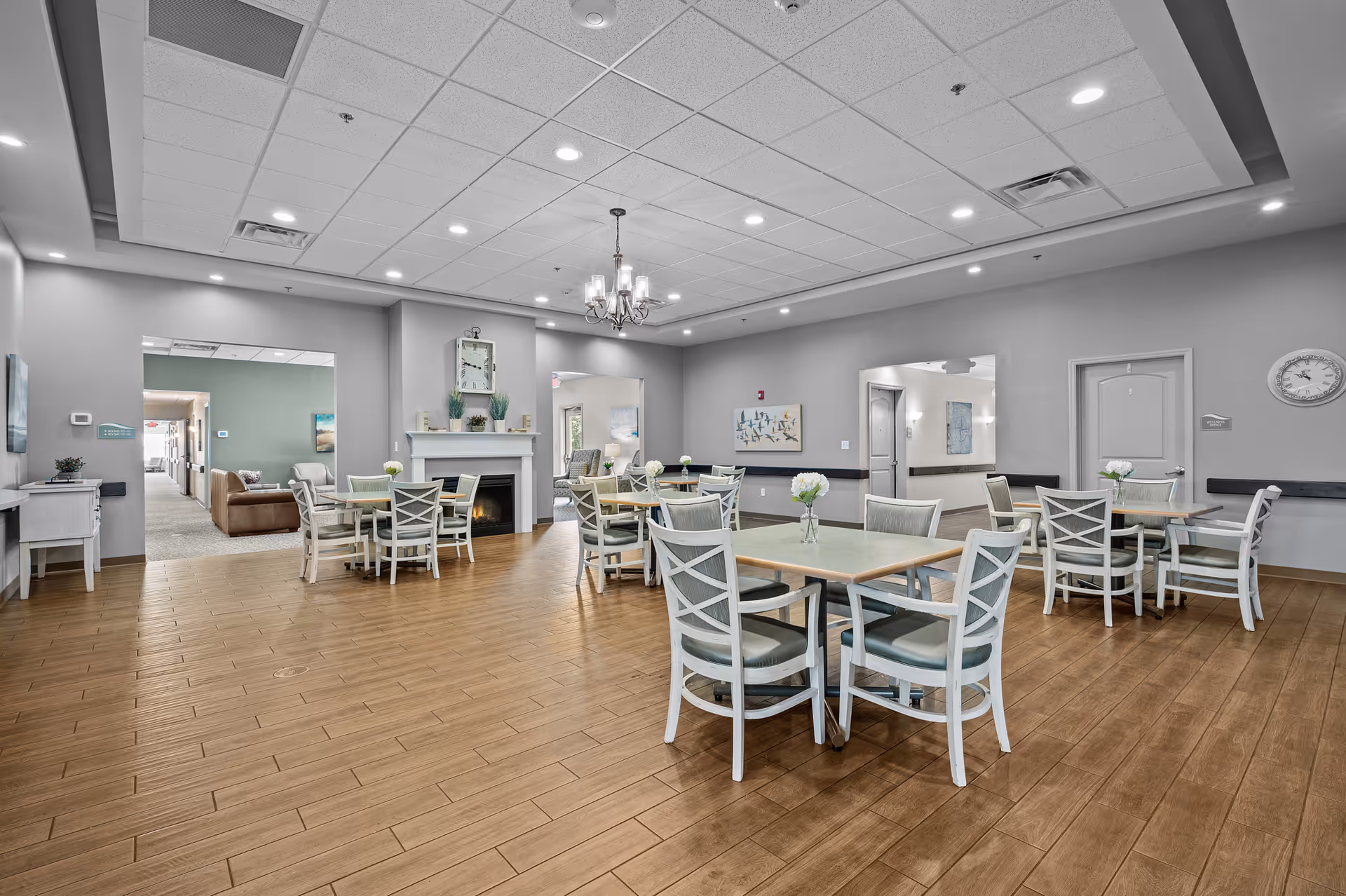 A spacious senior living dining room with several square tables and chairs arranged neatly. The room features wood-look flooring, a white fireplace with a clock and plants on the mantel, and soft gray walls. There is a chandelier hanging from the ceiling and multiple recessed lights. Open doorways lead to other rooms, including a lounge area with sofas.