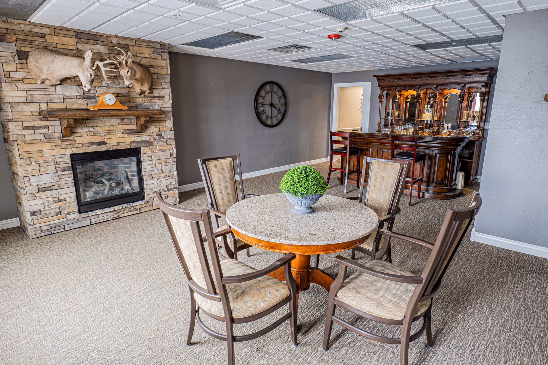 A cozy common area with a round table surrounded by four cushioned chairs, a stone fireplace with mounted deer heads above it, and a wooden bar with bar stools in the background. A decorative clock is mounted on the wall, and a small green plant centerpiece is on the table.