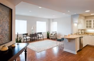 A bright and spacious interior living area featuring a kitchen with white cabinetry and a breakfast bar with stools. The living room area has a large rug, two chairs, a small table, and large windows allowing natural light to fill the space. Hardwood floors and recessed lighting complete the modern and cozy atmosphere.
