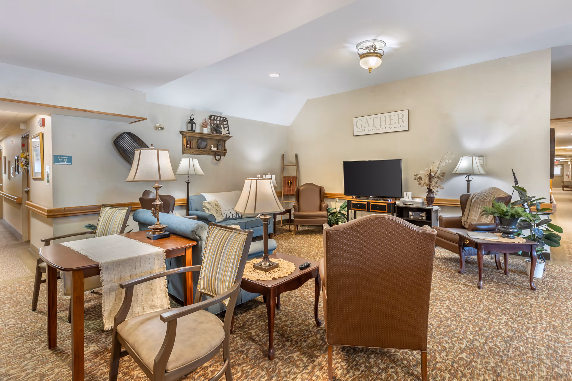 A cozy living room area in a senior living facility with multiple chairs, a sofa, several table lamps, a TV on a stand, decorative plants, and wall decor including a sign that says 'GATHER'. The room has carpeted flooring and soft lighting, with a hallway visible in the background.