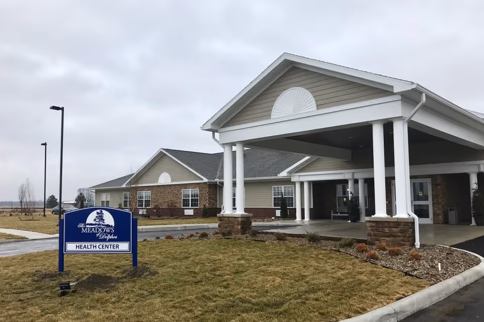 Exterior view of The Meadows of Delphos Health Center building with a covered entrance supported by white columns, a landscaped area with grass and small plants, and a blue sign in the foreground displaying the facility name and 'Health Center'. The sky is overcast.