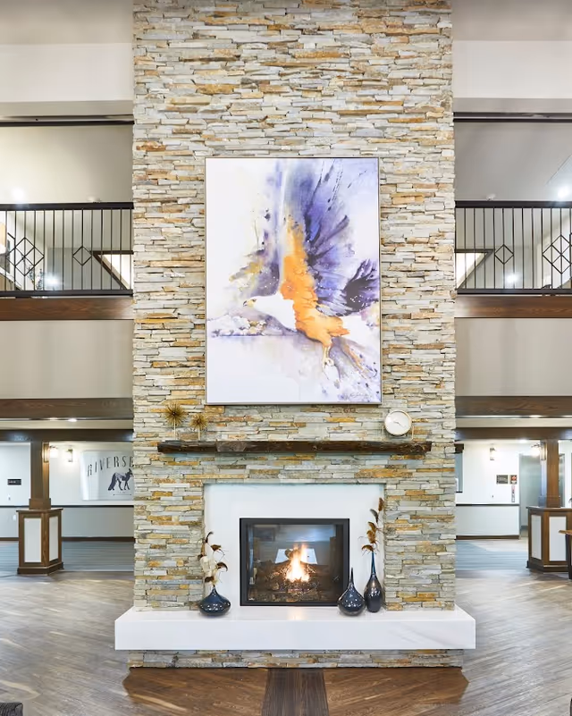 A tall stone fireplace with a lit fire in the center, decorated with three vases holding dried plants on the hearth and a wooden mantel holding decorative items including a clock. Above the mantel is a large colorful painting of a bird in flight. The fireplace is situated in a spacious interior with wooden railings on the upper level and light-colored walls.