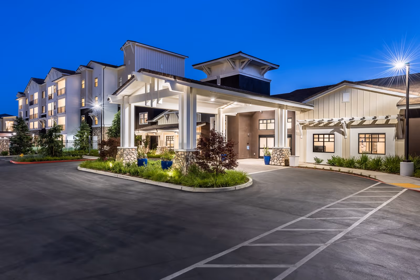 Exterior view of Revel Lodi senior living facility at dusk, showing a well-lit entrance with a covered drop-off area, landscaped greenery, and a multi-story building in the background under a clear blue sky.