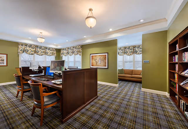 A well-lit interior room with green walls and plaid carpet featuring a wooden reception desk with two chairs and computer monitors. There is a bookshelf filled with books on the right side and a cozy seating area with a brown couch near windows with floral valances. A framed picture hangs on the wall behind the desk.
