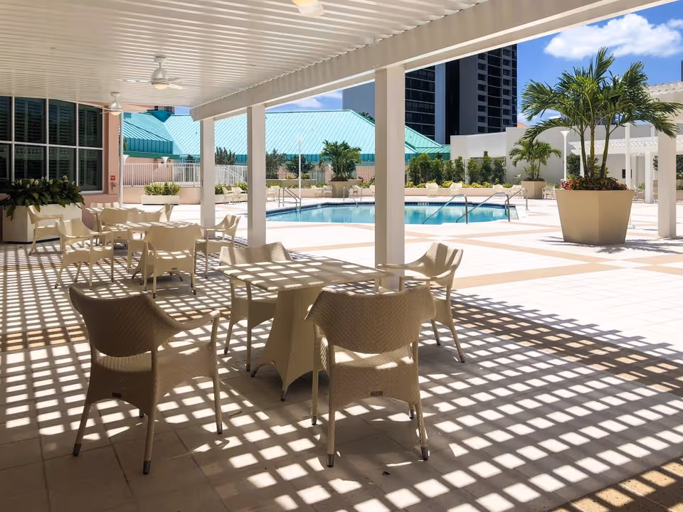 Outdoor patio area with several beige chairs and tables under a white pergola casting grid-like shadows on the tiled floor. In the background, there is a swimming pool surrounded by potted plants and buildings with a clear blue sky above.